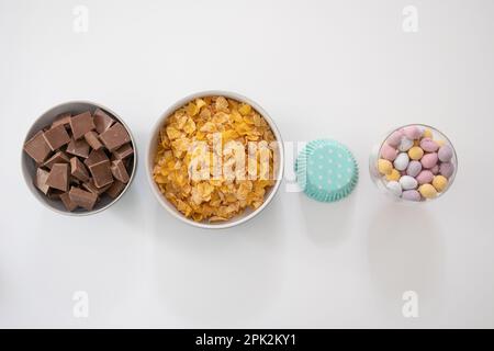 Zutaten in Schüsseln zum Herstellen von Schokoladennestern – Cornflakes, Schokoladenstücke, Mini-Eier und Cupcake-Kästen (Ostern) Stockfoto