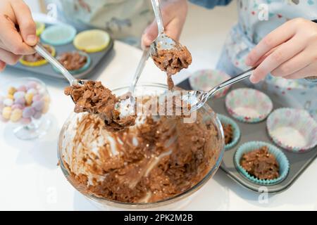 Zwei Kinder schöpfen Schokoladenmischung aus Maisflake und machen zu Ostern Schokoladennester aus Mini-Eiern Stockfoto