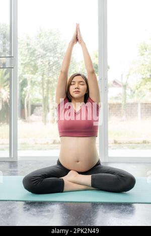 Glückliche junge, schwangere Asiatin in Fitnesskleidung, die während ihres Yoga-Trainings in ihrem Heim li die Beine kreuzt und sich die Arme hochstreckt Stockfoto