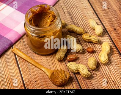 Becher mit cremiger Erdnussbutter auf dem Tisch und in Holzlöffel und Erdnüsse in Schalen, die auf dem Tisch verstreut sind Stockfoto