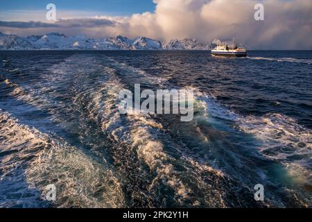 Wellen hinter Boot, Schiff und schneebedeckten Berg am Horizont mit dramatischem Himmel und Wolken, beleuchtet von untergehender Sonne. Lofoten, Norwegen. Stockfoto