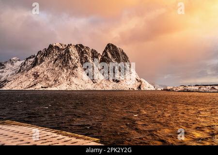Wunderschöner Sonnenaufgang über dem Meer und schneebedeckte felsige Berge, stimmungsvolle Atmosphäre in Sakrisoy, Lofoten, Norwegen. Stockfoto