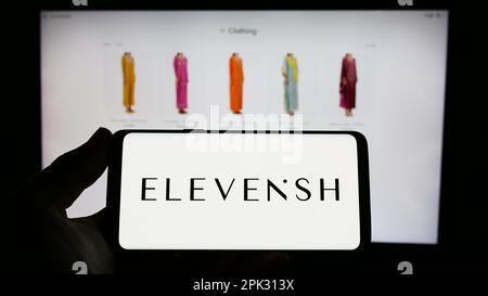 Person, die ein Smartphone mit dem Logo des eCommerce-Unternehmens ELEVENiSH auf dem Bildschirm vor der Website hält. Konzentrieren Sie sich auf das Display des Telefons. Stockfoto