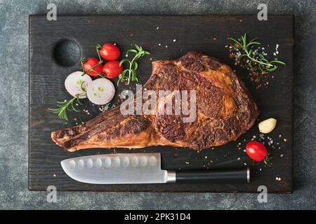 Tomahawk-Steak. Gegrilltes Tomahawk-Rindersteak mit gebackenen Kirschtomaten, Kräutern und Salz auf altem Holzhintergrund. Grillvorgang wird vorbereitet. Draufsicht Stockfoto