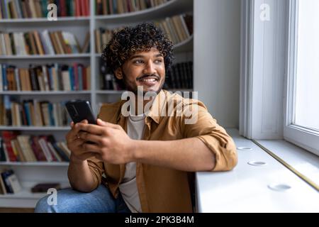 Nahaufnahme eines jungen erfolgreichen hispanischen Schülers, Mann mit lockigem Haar, der lächelnd aus dem Fenster schaut und glücklich ist, Teenager, der in der Bibliothek der Universität in der Hand sitzt. Stockfoto