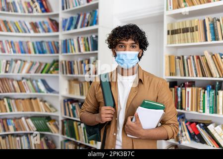 Porträt eines jungen männlichen Studenten in einer Bibliothek, der an einer Universität studiert, hispanischer Mann mit lockigem Haar, der auf die Kamera schaut, die Bücher in seinen Händen hält, der Kerl trägt eine schützende Gesichtsmaske. Stockfoto