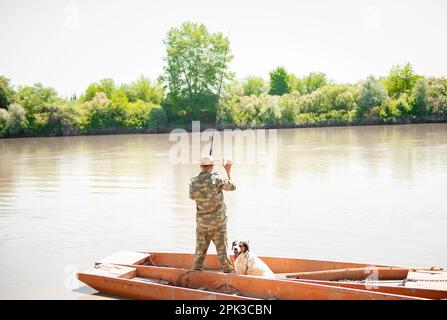 Starker männlicher Fischer in Camo-Kleidung, steht in einem alten Holzboot mit Stange, schwingt an sonnigen Tagen. Stockfoto