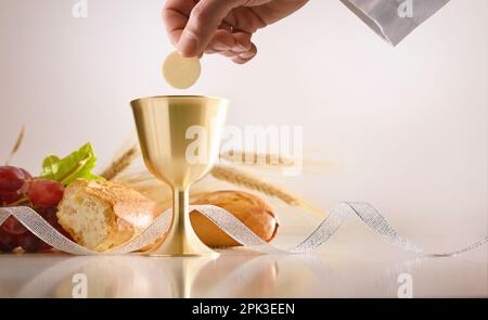 Eucharist mit der Hand des Priesters, der einen Wirt und eine Tasse geweihten Weins auf einem Tisch mit Brot und Trauben hält und einen isolierten Hintergrund hat. Vorderansicht. Stockfoto