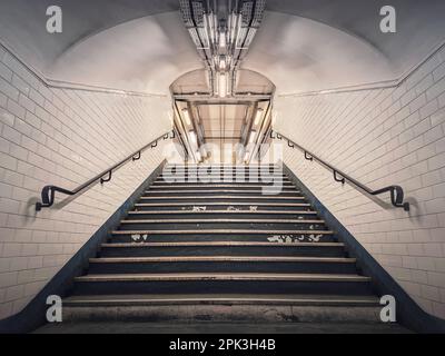 U-Bahn-Korridor mit weißen Fliesen an den Wänden und leuchtenden Lichtern über einem Treppengang. Symmetrische unterirdische Treppe Stockfoto