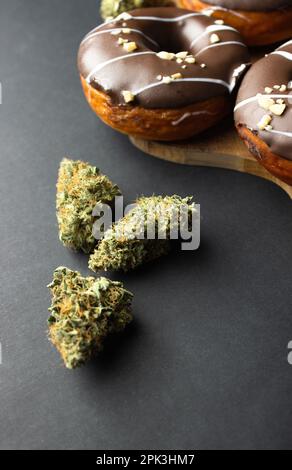 Trockene Knospen von medizinischem Marihuana aus der Nähe mit Donuts bedeckt mit Schokoladeneis, mit Nusskrümeln bestreut. Auf schwarzem Hintergrund Stockfoto