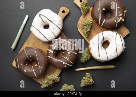 Donuts bedeckt mit Schokolade und weißer Glasur liegen auf Holzbrettern zwischen trockenen Knospen von medizinischem Marihuana, neben zwei Gelenken. Auf schwarzem Hintergrund, fl Stockfoto