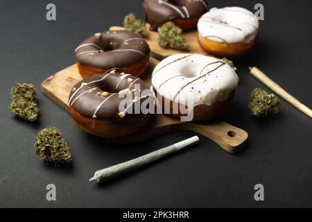 Donuts bedeckt mit Schokolade und weißer Glasur liegen auf Holzbrettern zwischen trockenen Knospen von medizinischem Marihuana, neben zwei Gelenken. Auf schwarzem Hintergrund Stockfoto