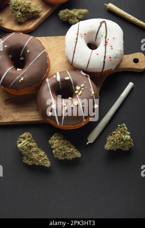 Donuts bedeckt mit Schokolade und weißer Glasur liegen auf Holzbrettern zwischen trockenen Knospen von medizinischem Marihuana, neben zwei Gelenken. Auf schwarzem Hintergrund, fl Stockfoto