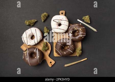 Donuts bedeckt mit Schokolade und weißer Glasur liegen auf Holzbrettern zwischen trockenen Knospen von medizinischem Marihuana, neben zwei Gelenken. Auf schwarzem Hintergrund, fl Stockfoto