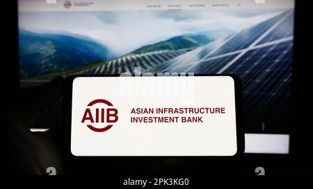 Person, die ein Smartphone mit dem Logo der Asiatischen Infrastruktur-Investitionsbank (AIIB) auf dem Bildschirm vor der Website hält. Konzentrieren Sie sich auf das Display des Telefons. Stockfoto