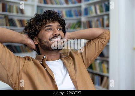 Ein erfolgreicher Student mit den Händen hinter dem Kopf, der sich tagsüber ausruht, träumt vom Studium an einem renommierten Universitätscollege, einem hispanischen Mann mit lockigem Haar, der in der Bibliothek geschlossen wird. Stockfoto
