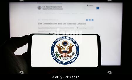 Person, die ein Mobiltelefon mit dem Logo der Kommission für Chancengleichheit am Arbeitsplatz (EEOC) auf dem Bildschirm vor der Webseite hält. Konzentrieren Sie sich auf das Display des Telefons. Stockfoto