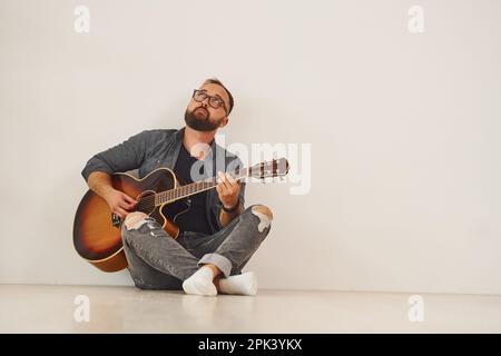 Er sitzt an der Wand. Mann in lässiger Kleidung und mit Akustikgitarre ist drinnen Stockfoto