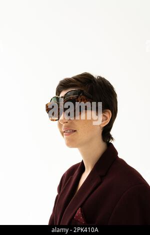 Brünette Frau in brauner Jacke, die mehrere trendige Sonnenbrillen trägt und auf weißem Hintergrund lächelt, Stockbild Stockfoto