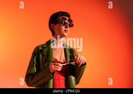 Trendige Frau in grüner Lederjacke posiert in mehreren Sonnenbrillen und blickt auf orangefarbenen Hintergrund weg, Stockbild Stockfoto