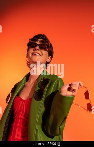 Aufregende und modische Frau in grüner Lederjacke mit verschiedenen Sonnenbrillen auf orangefarbenem Hintergrund, Stockbild Stockfoto