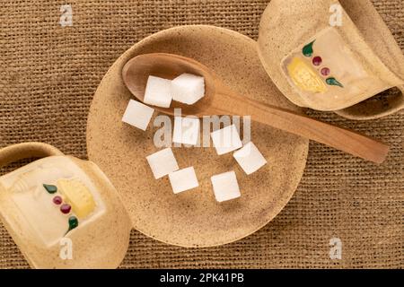 Mehrere weiße Zuckerwürfel mit Keramikuntertasse, Tassen, Holzlöffel auf Jutetuch, Makro, Draufsicht. Stockfoto