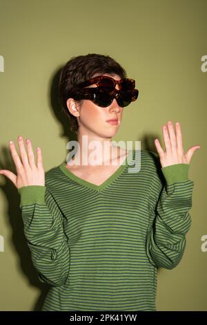 Brünette Frau in gestreiftem Pullover, die mehrere trendige Sonnenbrillen auf grünem Hintergrund trägt, Stockbild Stockfoto