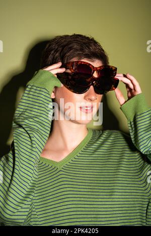 Junge Frau in trendy gestreiftem Pullover, trägt mehrere Sonnenbrillen auf grünem Hintergrund, Stockbild Stockfoto