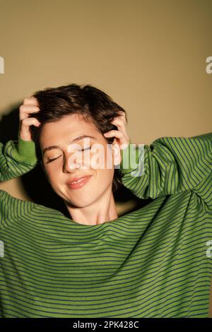 Zufriedene Frau in gestreiftem Pullover, die kurze braune Haare berührt und mit geschlossenen Augen auf grünem Hintergrund lächelt, Stockbild Stockfoto