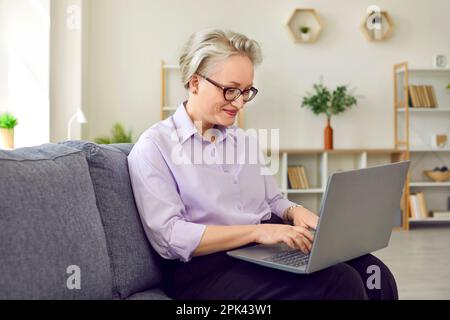 Eine reife Frau mit Brille arbeitet an einem Laptop und sitzt auf dem Sofa in ihrem gemütlichen Büro. Stockfoto