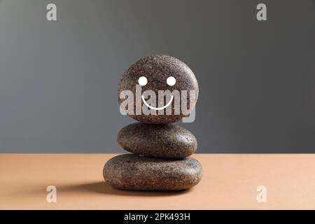 Steinstapel mit gemaltem Gesicht auf beigefarbenem Tisch vor grauem Hintergrund. Zen-Konzept Stockfoto