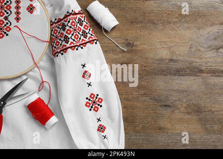 Hemd mit roter Stickerei im Ring, Schere und Fäden auf einem Holztisch, flach liegend. Ukrainische Nationalkleidung Stockfoto