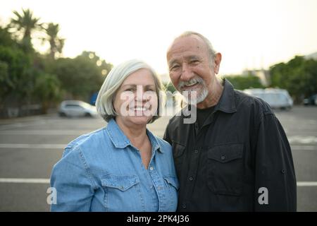 Porträt eines positiven älteren Ehepaars mit grauen Haaren, die lächeln und in die Kamera schauen, während man an sonnigen Tagen auf einer unscharfen Straße steht Stockfoto