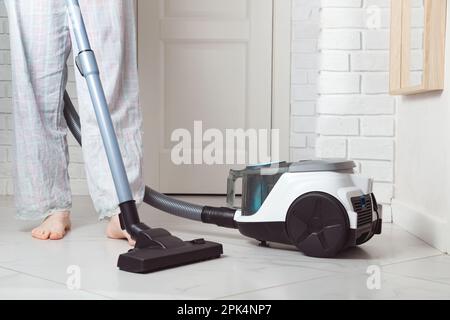 Barfußfrau reinigt Keramikfliesenboden mit Staubsauger im modernen weißen Innenraum. Hausarbeiten erledigen und die Wohnung putzen Stockfoto