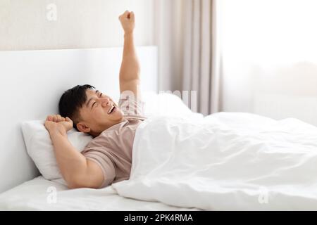 Positiver asiatischer Mann liegt im Bett und streckt sich die Arme Stockfoto