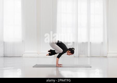 Seitenansicht einer charmanten Frau, die bakasana während der Yoga-Meditation in einem hellen Fitnessstudio macht. Das Konzept der Armfestigkeit und -Ausdauer, -Flexibilität und -Balance Stockfoto