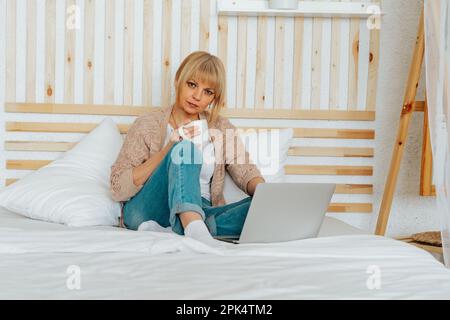 Fernunterricht, Online-Ausbildung und -Arbeit. Entspannen Sie sich, wenn eine Erwachsene Frau am Laptop tippt, auf dem Bett sitzt und Kaffee trinkt. Lächelnder Senior mit Notizbuch Stockfoto
