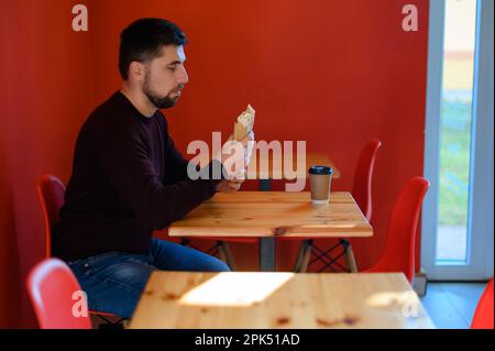 Ein Mann mit Bart sitzt in einem Fast-Food-Restaurant und hält ein Schawarma in der Hand. Er hat ein Glas Kaffee, eine rote Wand und einen Holztisch. Stockfoto