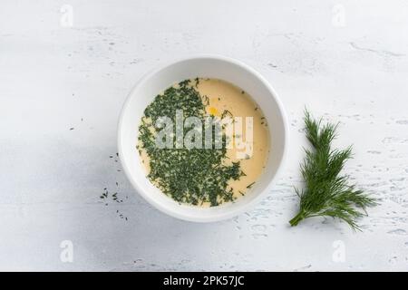 Eine weiße Schüssel mit einem geschlagenen Ei, Milch und Grün auf hellblauem Hintergrund, Draufsicht. Omelette kochen, hausgemachte gesunde Speisen Stockfoto