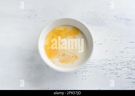 Eine weiße Schüssel mit einem geschlagenen Ei und Milch auf hellblauem Hintergrund, Draufsicht. Omelette kochen, hausgemachte gesunde Speisen Stockfoto