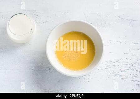 Eine weiße Schüssel mit einem geschlagenen Ei und einer Kanne Milch auf hellblauem Hintergrund, Draufsicht. Omelette kochen, hausgemachte gesunde Speisen Stockfoto