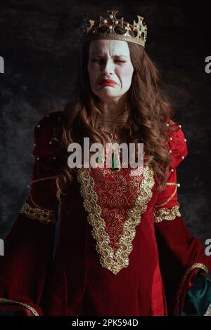 Traurige mittelalterliche Königin in rotem Kleid mit weißem Make-up und Krone auf dunkelgrauem Hintergrund. Stockfoto