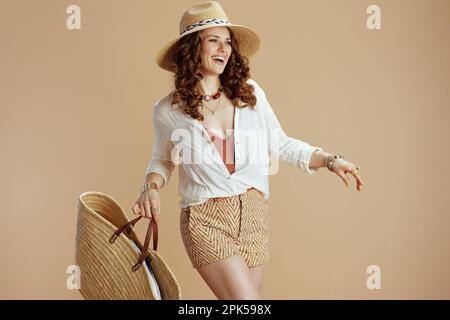 Strandurlaub. Lächelnde elegante Frau in weißer Bluse und Shorts, isoliert auf beigefarbenem Hintergrund mit Strohtasche und Strohhut. Stockfoto