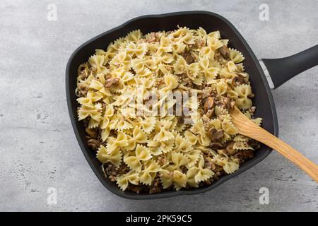 Traditionelles jüdisches Gericht Kasha Varnishkes: Buchweizen, Champignon Pilze, gebratene Zwiebeln, Farfalle Pasta in einer antihaftbeschichteten Pfanne auf einem grauen strukturierten B Stockfoto