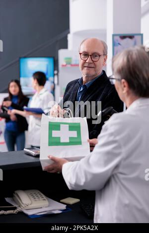 Apotheke, die eine alte Drogerie online bestellt, an der Kasse hält Papiertüte mit dem Kauf des Kunden in der Hand. Älterer Brillenkäufer beim Arztbesuch und bei der Einnahme von Medikamenten an der Kasse Stockfoto