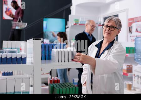 Portrait des älteren Apothekers steht neben Apothekenregalen Pillen Pakete helfen Kunden, Arbeiter bietet medizinischen Service und Unterstützung. Verschiedene Menschen in der Apotheke, die Vitamine kaufen Stockfoto