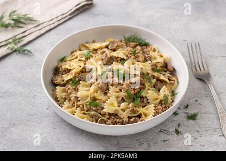 Traditionelles jüdisches Gericht Kasha Varnishkes: Buchweizen, Pasta, Champignon-Pilze, gebratene Zwiebeln mit Kräutern auf grauem, strukturiertem Hintergrund, vegane Speisen, t Stockfoto