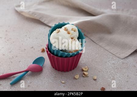 Vanilleeisschaufeln oder Kugeln mit Nüssen in bunten Bambusbechern auf beigefarbenem strukturiertem Hintergrund Stockfoto