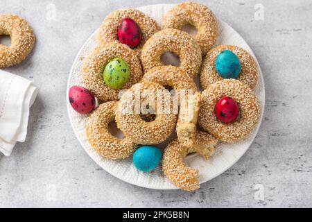 Weißer Teller mit traditionellen griechischen Osterkeksen Koulourakya oder Koulourya Shortbread Ringe mit Sesamsamen, dekoriert mit farbigen Eiern auf einem grauen T Stockfoto