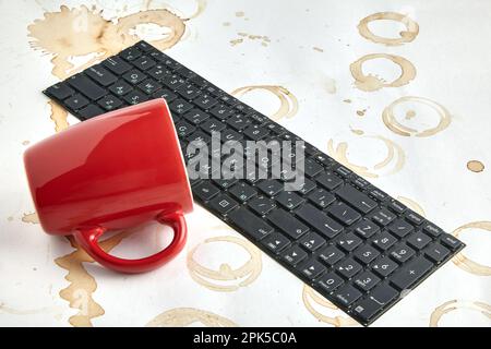 Die rote Kaffeetasse wurde auf einer Computertastatur auf einem Tisch mit Kaffeeflecken umgeworfen. Tastatur gefüllt mit Kaffeegetränk Stockfoto